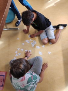 Enfants sur sol beige jouant avec des cartes triangulaires, un adulte se tenant proche.