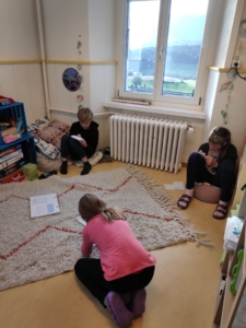 Trois enfants lisent dans une pièce lumineuse avec radiateur, fenêtre et étagère de jeux.